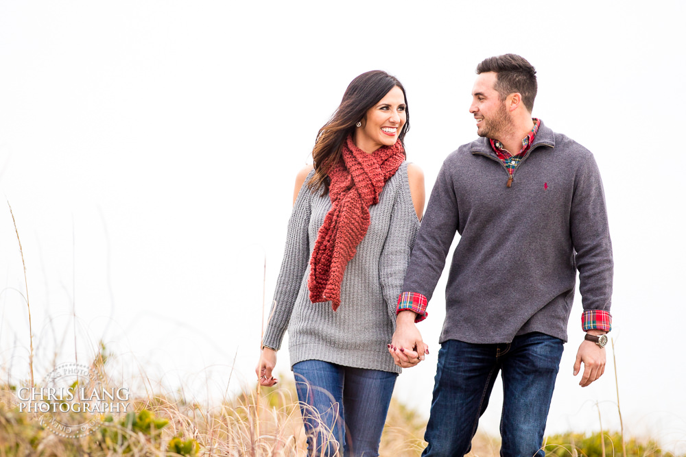 image of  a couple walking on the beach holding hands - Fall coloer beach outfit ideas - Wrightsville Beach Engagement Photography - Wrightsville Beach Photographers - Engagement Photographer -  Chris Lang Photography - Lifestyle Engagement Photo