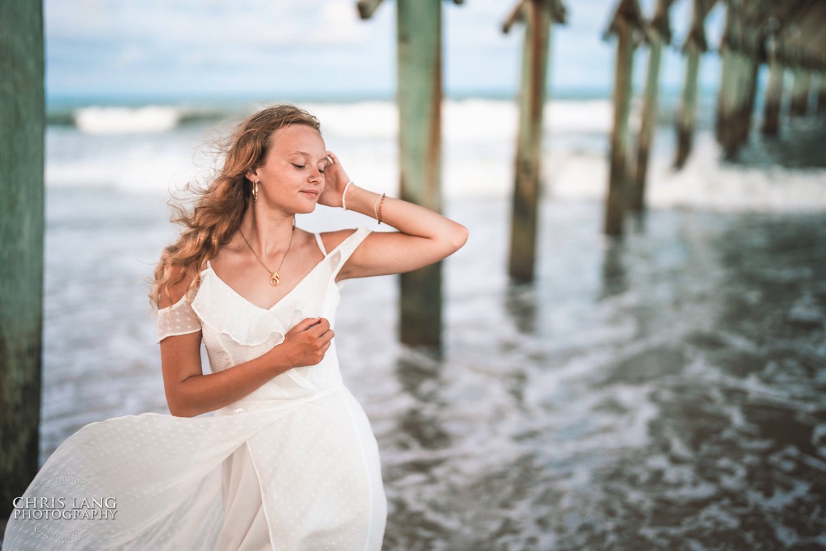 photo of highschool senior girl posing on the beach  under a pier wearing a white dress - senior portrait ideas - Wrightsville Beach High School Senior Photography - Wrightsville Beach Photographers - Beach Senior Portrait  Photos- Chris Lang Photography  