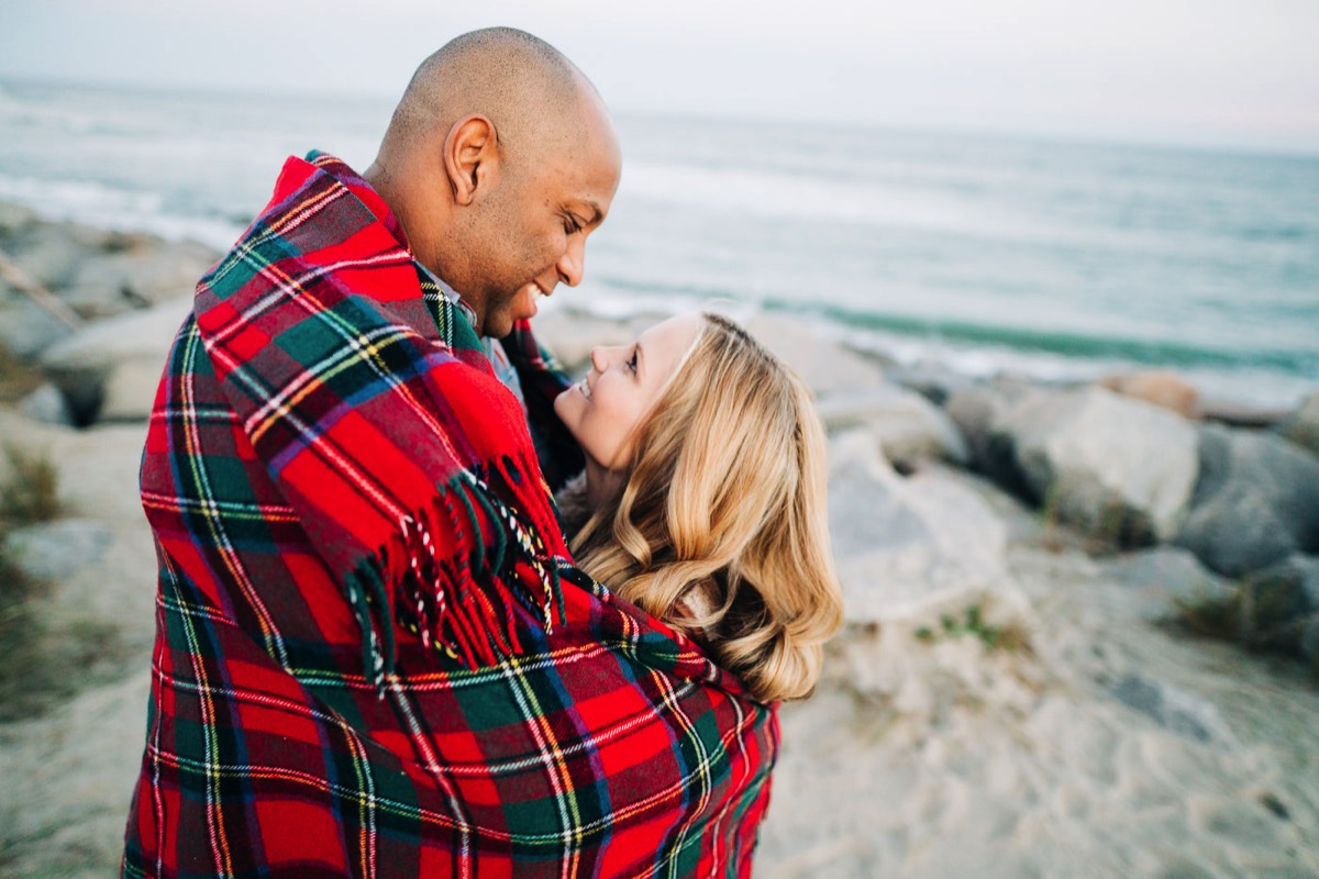 Couple wrapped in a red blanket on the beach - Fort Fisher North Carolina -  Engagement Photography - Wedding Ideas - Popular engagement photography locations - Lifestyle engagement photo -  Ft Fisher engagement photographers - Engagement session ideas - New trends in engagement photography - Chris Lang Photography - Engagement photos  